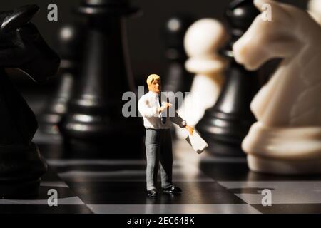 Un professeur miniature explique les règles du jeu d'échecs à bord. Concept d'analyse de stratégie d'échecs. Figurines d'échecs noir et blanc et homme d'affaires. Apprentissage des échecs Banque D'Images