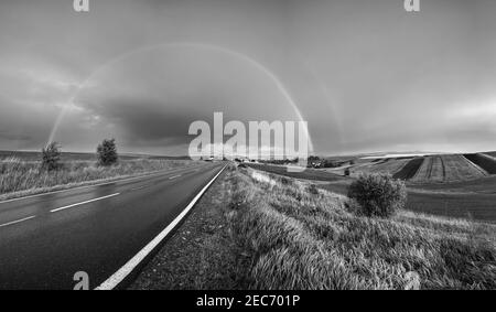 Niveaux de gris. Champ de graines de colza et de petites terres agricoles après la pluie vue en soirée, ciel nuageux avant coucher de soleil avec arc-en-ciel et collines rurales. Saisonnier, météo, Banque D'Images