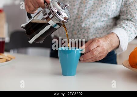 Main de l'homme versant du café aromatique dans une tasse bleue de la presse française sur une table en bois pendant le petit déjeuner. Personne âgée le matin buvant un espresso brun frais provenant d'une tasse d'époque, filtre relax rafraîchissement Banque D'Images