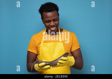 Jeune homme africain en tablier jaune avec poisson de saumon entier. Il contrôle la qualité des aliments sur le marché. Prise de vue en studio Banque D'Images