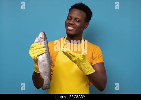 Jeune homme africain en tablier jaune avec poisson de saumon entier. Il contrôle la qualité des aliments sur le marché. Prise de vue en studio Banque D'Images