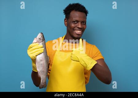 Jeune homme africain en tablier jaune avec poisson de saumon entier. Il contrôle la qualité des aliments sur le marché. Prise de vue en studio Banque D'Images