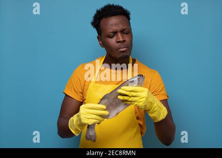 Jeune homme africain en tablier jaune avec poisson de saumon entier. Il contrôle la qualité des aliments sur le marché. Prise de vue en studio Banque D'Images