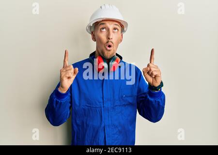 Homme chauve avec la barbe portant le constructeur combinaison uniforme et casque de sécurité stupéfait et surpris regardant vers le haut et pointant avec les doigts et les bras relevés. Banque D'Images