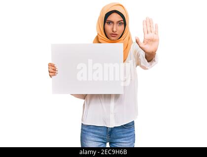 Une jeune femme arabe brunette portant le hijab islamique traditionnel tenant une bannière à main ouverte faisant signe d'arrêt avec une expression sérieuse et confiante, defen Banque D'Images