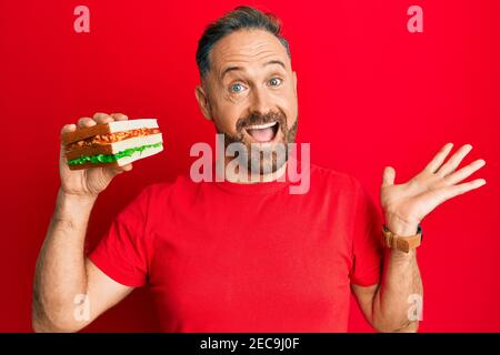 Beau homme d'âge moyen mangeant un sandwich célébrant la réussite avec heureux sourire et expression du gagnant avec la main levée Banque D'Images