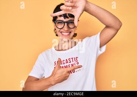 Belle femme brunettte portant des commentaires sarcastique chargement t-shirt sourire faire un cadre avec les mains et les doigts avec le visage heureux. Créativité et photo Banque D'Images