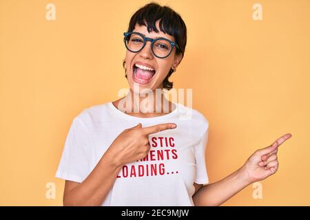 Belle femme brunettte portant des commentaires sarcastiques chargement t-shirt sourire et regarder la caméra pointant avec deux mains et deux doigts sur le côté Banque D'Images