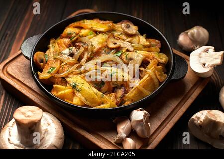 Pommes de terre frites maison, avec oignons, champignons et ail, dans une poêle, sur une planche en bois Banque D'Images