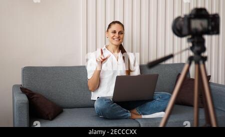 Une femme blogueuse souriante enregistre une nouvelle vidéo. Elle est assise sur le canapé à la maison et tient un ordinateur portable. Concept positif de blog d'affaires. Banque D'Images