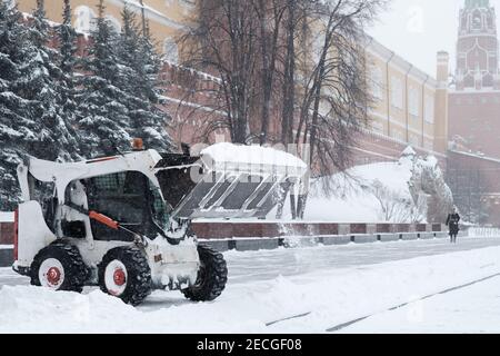 Un petit bobcat de pelle-chargeuse enlève la neige du trottoir près des murs du Kremlin lors d'une forte chute de neige. Des flocons de neige volent dans l'air. Banque D'Images