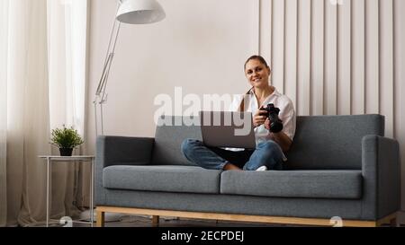 Jeune femme heureuse avec appareil photo utilisant un ordinateur portable à la maison. Le photographe tient l'appareil photo et sourit. Banque D'Images