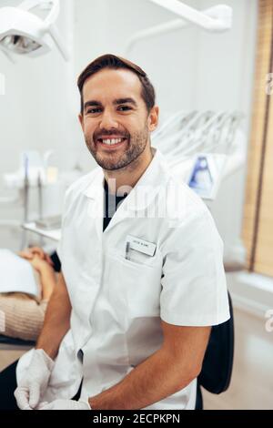 Portrait d'un expert dentaire confiant dans sa clinique. Un dentiste de sexe masculin portant un blouse de laboratoire, regardant l'appareil photo et souriant. Banque D'Images