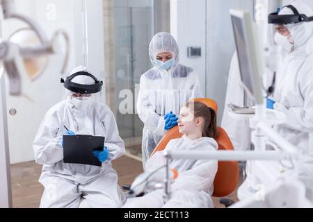 Petite fille souriant chez un dentiste pédiatrique assis sur une chaise dentaire au cours de la vérification dentaire. Stomatologiste pendant la covid19 port de l'epi costume faire les dents procédure de l'enfant assis sur une chaise. Banque D'Images