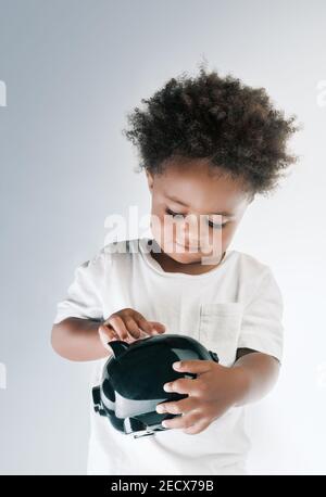 Portrait d'un enfant afro-américain de Nice mettant des pièces dans la boîte à monnaie en forme de cochon isolée sur fond gris blanc. Investir dans l'avenir Banque D'Images