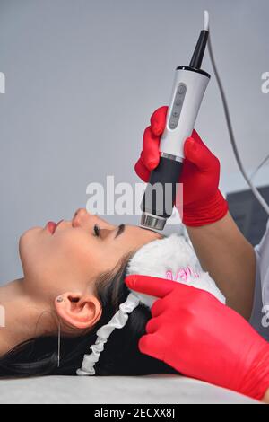 jeune femme maître de l'enlèvement de cheveux de laser, laser à part, femme sourit, elle est en gants roses. Section de cosmétologie épilation. Mains en gants roses mak Banque D'Images