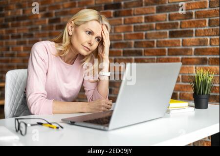 Découragé femme d'âge moyen assis à l'ordinateur portable, ne sait pas quoi faire, au bureau, regardant l'écran de l'ordinateur portable, inquiet et fatigué, un entrepreneur Banque D'Images