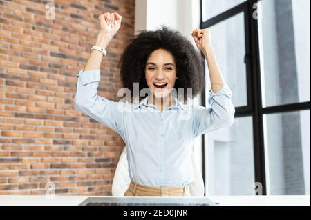 Bonne jeune femme d'affaires avec les mains courtes et bouclés, assise devant l'ordinateur, avec un grand sourire, se réjouit de son succès, a obtenu un nouvel emploi, a reçu Banque D'Images