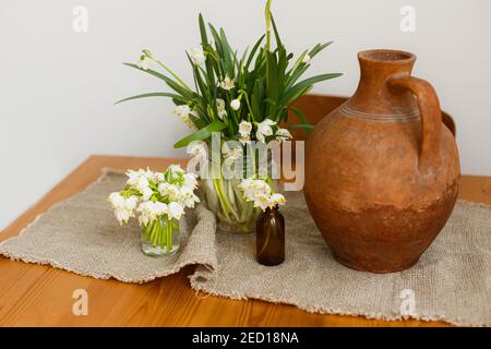 Fleurs printanières dans des bouteilles en verre et vase en terre cuite sur une table rustique en bois avec linge de lit dans la chambre. La vie rurale encore. Bonjour printemps. Jardinage et bo Banque D'Images