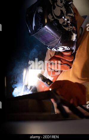 Employé masculin méconnaissable dans des gants de protection et un casque par soudage machine lors de travaux dans un atelier sombre Banque D'Images