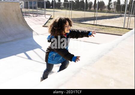 Vue latérale de l'enfant ethnique positif avec la course à pied à la coiffure afro prenez la rampe tout en vous amusant au skate park le week-end jour ensoleillé Banque D'Images