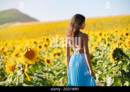 Vue arrière d'une femme ethnique mince et belle debout dans le champ avec des tournesols fleuris le jour ensoleillé en été regarder plus l'épaule Banque D'Images