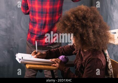 Rogné enseignant non reconnaissable écrivant l'exemple mathématique sur tableau noir pour le noir fille assise à un bureau en classe pendant la leçon à l'école Banque D'Images