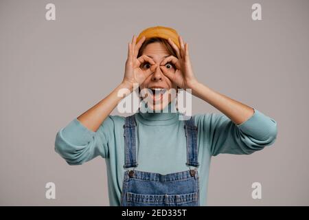 Photo de la jolie femelle hipster tend à regarder dans les verres. Blanc femme porte une combinaison en denim et chapeau isolé de couleur grise fond Banque D'Images