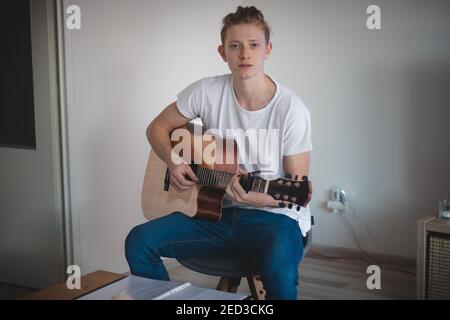 Portrait décontracté d'un jeune homme assis sur un tabouret bas dans une pièce éclairée jouant un instrument instrumental de guitare. La joie d'un nouveau passe-temps. La joie Banque D'Images