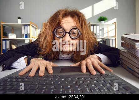Drôle fou regardant nerd dans les verres épais assis dans bureau et utilisation d'un ordinateur portable Banque D'Images
