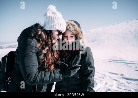 Deux amis dans la neige sourient tandis que l'un montre son téléphone cellulaire à l'autre. Banque D'Images