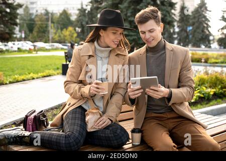 Jeune femme reposante regardant l'écran du smartphone tenu par son petit ami assis sur le banc et en discutant et café Banque D'Images