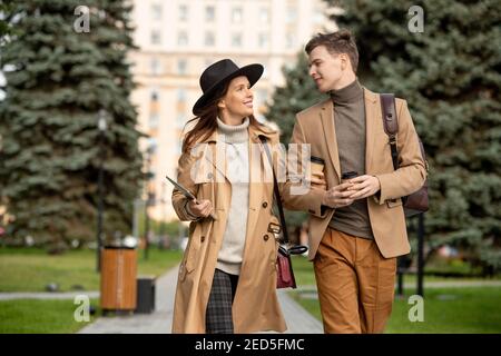 Joyeux jeune dates dans des vêtements décontractés beige parler et prendre le café en mouvement dans l'environnement urbain contre la construction de contemporain université Banque D'Images