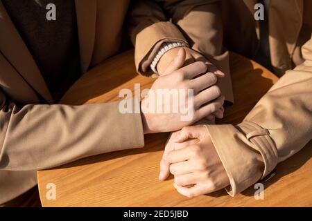 Mains d'un jeune homme et d'une femme affectueux en trenchcoats beige assis près d'une petite table ronde en bois l'une de l'autre par beau temps Banque D'Images