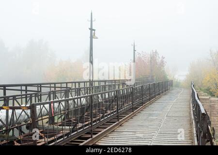 Perspective d'une longue route ou d'un pont entouré d'une clôture métallique, de tuyaux et d'arbres avec un feuillage jaune sur un matin d'automne brumeux et brumeux Banque D'Images