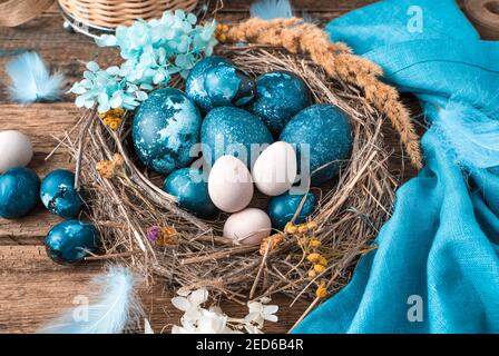 Œufs de Pâques bleu marbre dans un nid en osier avec plumes et fleurs à côté d'une serviette bleue et d'un panier d'œufs pastel. Banque D'Images