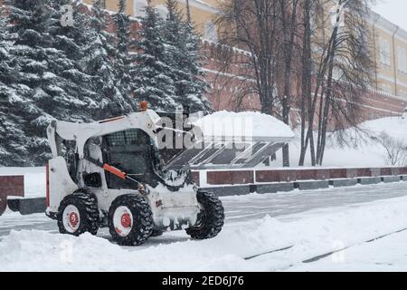 Un petit bobcat de pelle-chargeuse enlève la neige du trottoir près des murs du Kremlin lors d'une forte chute de neige. Des flocons de neige volent dans l'air. Banque D'Images