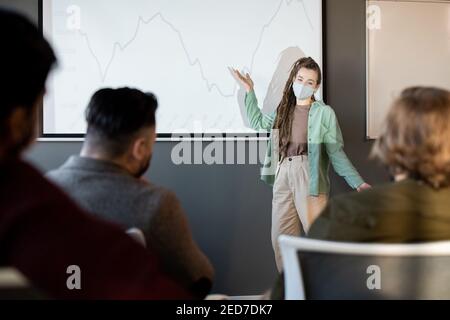 Jeune coach d'affaires confiant en vêtements décontractés et masque de protection pointant au graphique sur tableau blanc pendant la présentation pour les économistes Banque D'Images