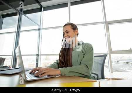 Bonne jeune femme d'affaires contemporaine en tenue décontractée avec un ordinateur portable assis par le lieu de travail contre une grande fenêtre dans un bureau à espace ouvert Banque D'Images