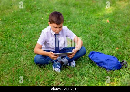 Un écolier sérieux est assis dans une chemise blanche et une cravate bleue sur l'herbe verte et regarde dans une tablette Banque D'Images