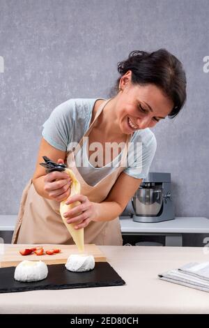 Le chef pâtissier de la jeune femme remplit les brownies de crème avec un sac de pâtisserie. Processus de fabrication du gâteau Anna Pavlova Banque D'Images