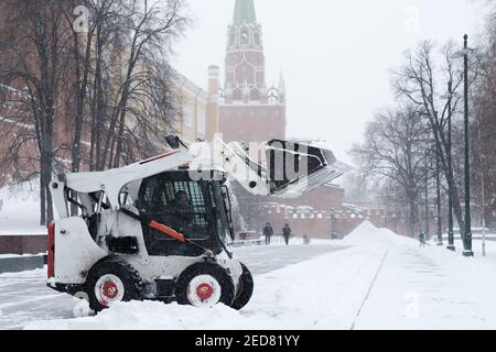 Un petit bobcat de pelle-chargeuse enlève la neige du trottoir près des murs du Kremlin lors d'une forte chute de neige. Des flocons de neige volent dans l'air. Banque D'Images