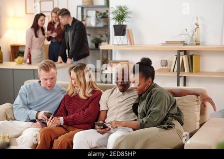 Deux jeunes couples interculturels en vêtements décontractés élégants assis à la rangée sur le canapé et discuter des photos sur les réseaux sociaux en mobile gadgets Banque D'Images