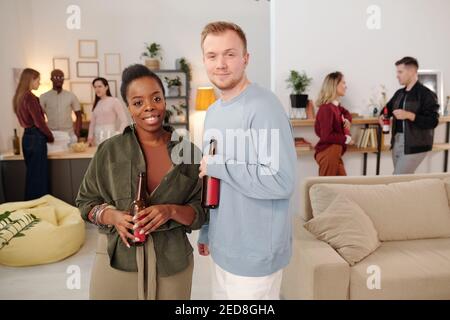 Joyeux jeune couple interculturel avec des bouteilles de bière regarder vous tout en profitant de la fête à la maison contre leurs amis discutant et prendre des boissons Banque D'Images