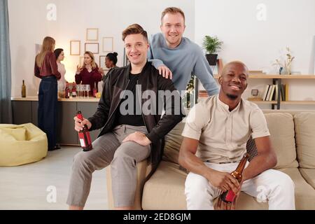 Joyeux jeunes hommes interculturels avec des bouteilles de bière regarder vous avec des sourires tout en appréciant la fête à la maison contre leurs petites amies Banque D'Images
