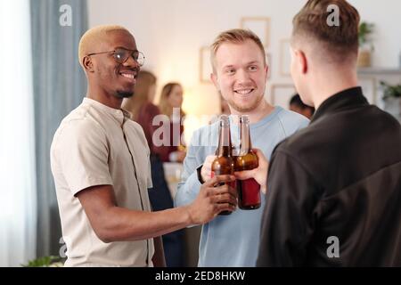 Joyeux jeunes hommes interculturels se clinquant avec des bouteilles de bière et se regarder les uns les autres avec des sourires crasseux tout en applaudisant à la fête à la maison Banque D'Images