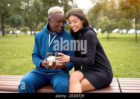 African american man proposes coffee asian woman Multiethnic friendship. Interracial relationship Young adult international couple drink hot beverage spending time together outdoor bench park Summer Stock Photo