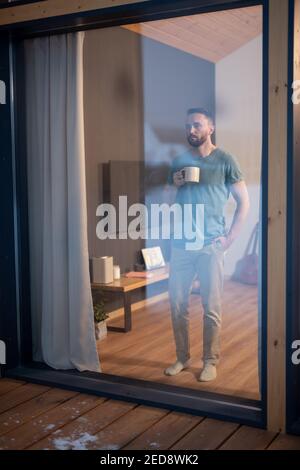 Jeune homme barbu en tenue décontractée tenant une tasse avec une boisson chaude et vue à travers la grande fenêtre tout en étant debout sur le plancher en bois dans la salle de séjour Banque D'Images