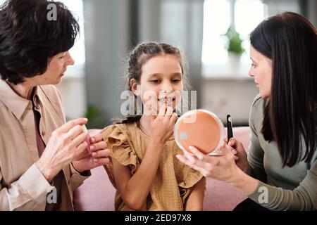 Jolie petite fille en robe appliquant un baume nutritif sur les lèvres tout en regardant dans le miroir tenu par la jeune femme brunette donnant ses conseils de beauté