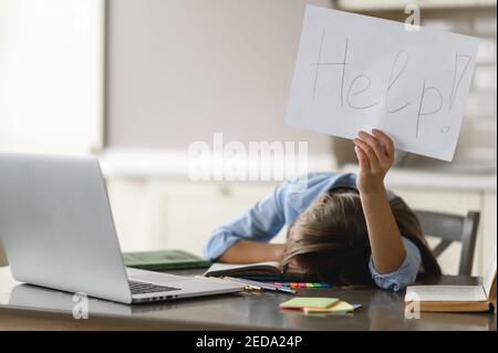 Formation à distance. La jeune fille est fatiguée d'étudier. Une écolière caucasienne frustrée, fatiguée d'étudier à la maison, se trouve à son bureau et tient une brochure avec l'aide d'inscription Banque D'Images
